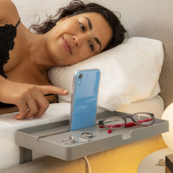 V0103325_0Prateleira Universal para Cama Bedten InnovaGoods – Organize e Tenha Tudo ao Alcance Mulher deitada confortavelmente usando a prateleira universal InnovaGoods para apoiar o telemóvel e objetos pessoais ao lado da cama.
